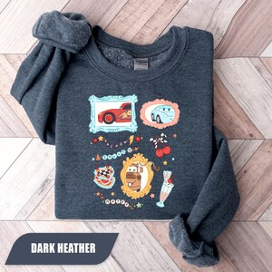 Puede incluir: Una sudadera de cuello redondo en gris jaspeado oscuro con un estampado gr&aacute;fico de personajes de la pel&iacute;cula Cars. El dise&ntilde;o incluye ilustraciones enmarcadas de Rayo McQueen, Sally y Mate, junto a iconos como un batido, cerezas y texto.