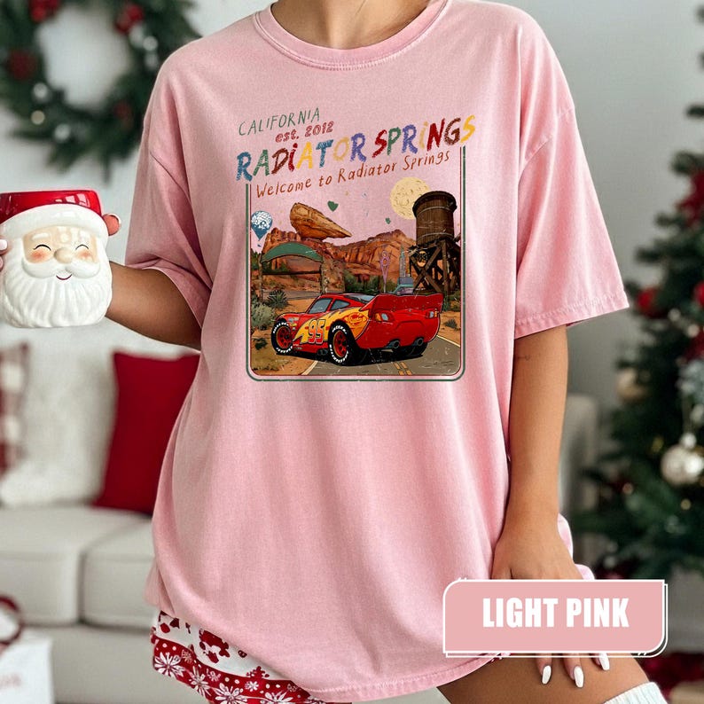 May include: A light pink t-shirt featuring a graphic print of a red race car on a desert road. The text reads California est. 2012 Radiator Springs Welcome to Radiator Springs. The design includes a water tower, rock formations, and a moon.