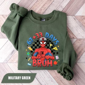 May include: Military green sweatshirt featuring a graphic of Spider-Man with the text "67+33 DAYS BRUH". The design includes a checkered background and various school-themed illustrations. The sweatshirt is made of a soft material.