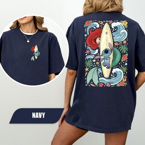 May include: A navy blue t-shirt featuring a small graphic on the front left chest and a large graphic on the back. Both graphics depict a blue alien character on a surfboard with a red-haired mermaid, ocean waves, tropical flowers, and a ukulele.