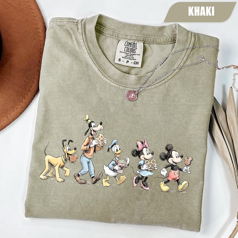 May include: A khaki-colored crew neck t-shirt featuring a graphic print of Pluto, Goofy, Donald Duck, Minnie Mouse, and Mickey Mouse eating snacks. The shirt has a Comfort Colors brand label inside the collar and a silver-toned necklace resting on it.