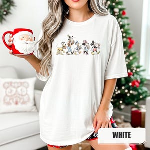 May include: A white t-shirt featuring a graphic print of Pluto, Goofy, Donald Duck, Minnie Mouse, and Mickey Mouse walking in a line. The shirt is paired with a red and white Santa Claus face mug with a handle on the left side of the ceramic vessel.