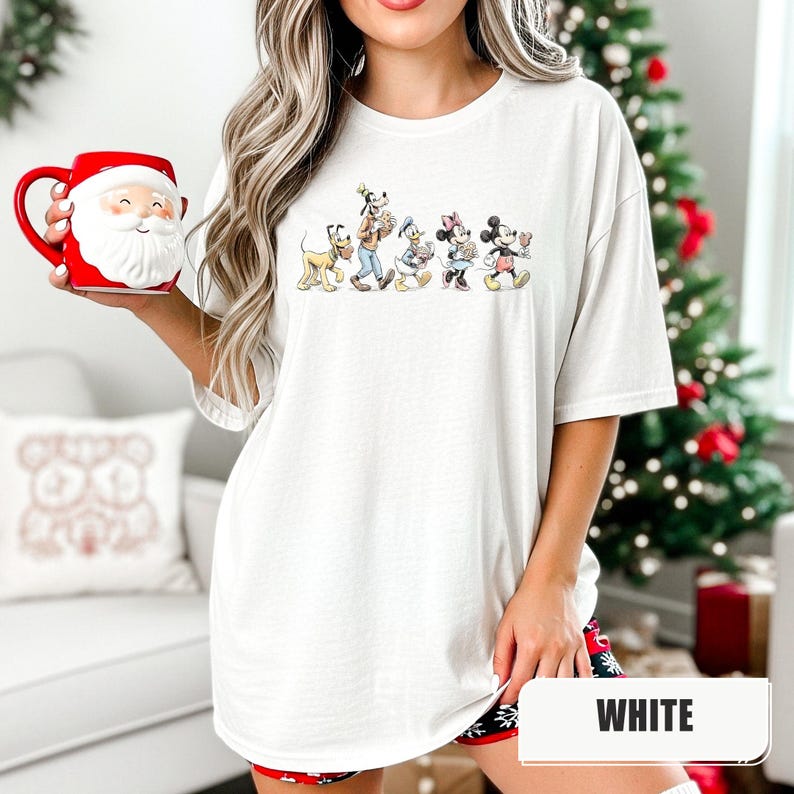 May include: A white t-shirt featuring a graphic print of Pluto, Goofy, Donald Duck, Minnie Mouse, and Mickey Mouse walking in a line. The shirt is paired with a red and white Santa Claus face mug with a handle on the left side of the ceramic vessel.