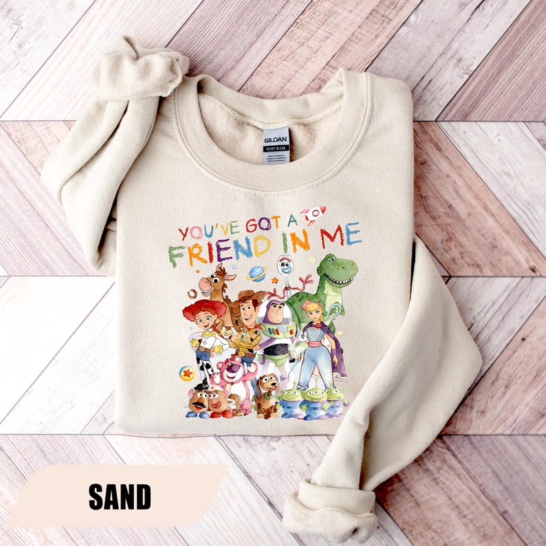 May include: A sand-colored crewneck sweatshirt featuring a colorful graphic print of various animated characters from the Toy Story franchise. The text "YOU'VE GOT A FRIEND IN ME" is printed in multi-colored letters above the character group.