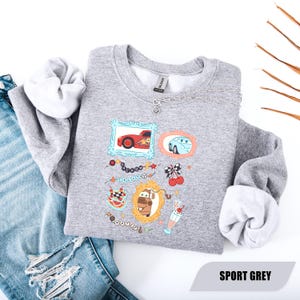 Puede incluir: Una sudadera gris deportivo de cuello redondo con un estampado gr&aacute;fico de tres marcos ornamentados que contienen coches de dibujos animados, incluyendo un coche de carreras rojo, uno azul y una gr&uacute;a marr&oacute;n. Incluye cerezas y un batido.