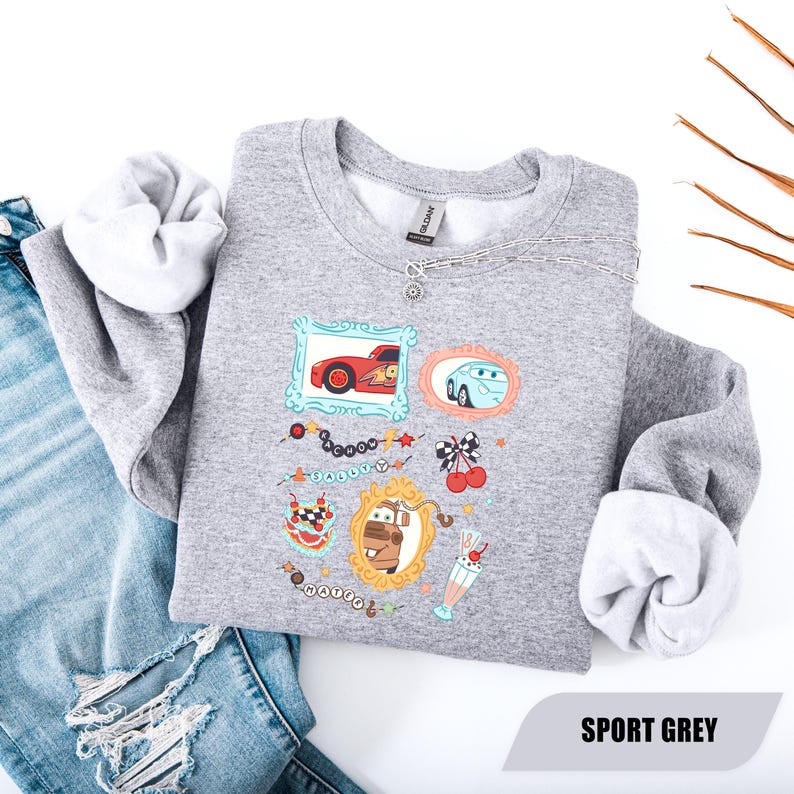 Puede incluir: Una sudadera gris deportivo de cuello redondo con un estampado gr&aacute;fico de tres marcos ornamentados que contienen coches de dibujos animados, incluyendo un coche de carreras rojo, uno azul y una gr&uacute;a marr&oacute;n. Incluye cerezas y un batido.