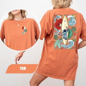 May include: An orange t-shirt featuring a small graphic on the front left chest and a large graphic on the back. The design depicts a blue alien character surfing on a surfboard alongside a mermaid, surrounded by tropical flowers, waves, and a ukulele.