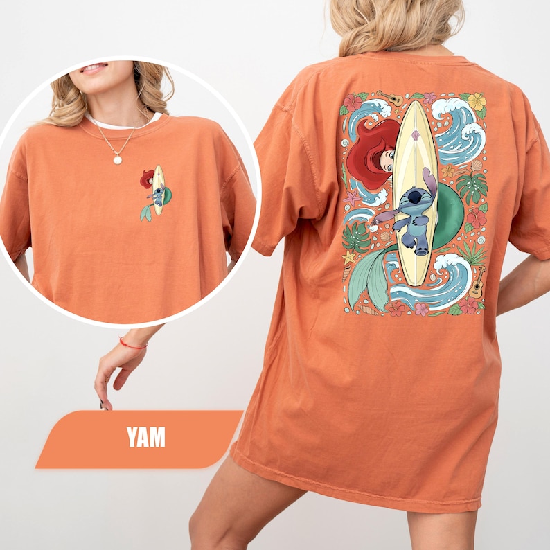 May include: An orange t-shirt featuring a small graphic on the front left chest and a large graphic on the back. The design depicts a blue alien character surfing on a surfboard alongside a mermaid, surrounded by tropical flowers, waves, and a ukulele.