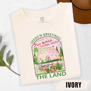 Könnte beinhalten: Elfenbeinfarbenes T-Shirt mit einer farbenfrohen Grafik mit einer geodätischen Kuppel, Figuren und dem Text "SEEDS'N GREETINGS From LIVING WITH THE LAND." Das Shirt hat einen Rundhalsausschnitt und kurze Ärmel. Die Marke Comfort Colors ist sichtbar.
