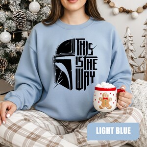 May include: Light blue sweatshirt featuring a black graphic of a helmet and the text "THIS IS THE WAY". The sweatshirt is a crew neck style. A white mug with a gingerbread man design is held in the hand.