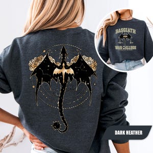May include: Dark heather gray sweatshirt featuring a black and gold dragon design on the back. The dragon is set against a constellation background with gold accents. The front of the sweatshirt has the text "BASGIATH WAR COLLEGE DRAGON RIDERS".