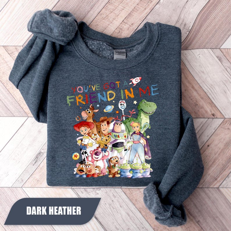 May include: A dark heather gray crewneck sweatshirt featuring a colorful graphic print of various animated toy characters. The text "YOU'VE GOT A FRIEND IN ME" is printed above the characters in a multi-colored, hand-drawn style font.