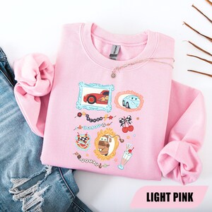 Puede incluir: Una sudadera de cuello redondo rosa claro con un estampado gr&aacute;fico de personajes de la pel&iacute;cula Cars. El dise&ntilde;o incluye ilustraciones enmarcadas de Rayo McQueen, Sally y Mate, junto con peque&ntilde;os iconos de cerezas, un batido y texto.
