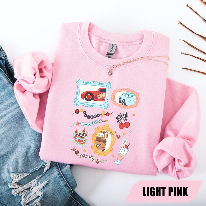 Puede incluir: Una sudadera de cuello redondo rosa claro con un estampado gr&aacute;fico de personajes de la pel&iacute;cula Cars. El dise&ntilde;o incluye ilustraciones enmarcadas de Rayo McQueen, Sally y Mate, junto con peque&ntilde;os iconos de cerezas, un batido y texto.