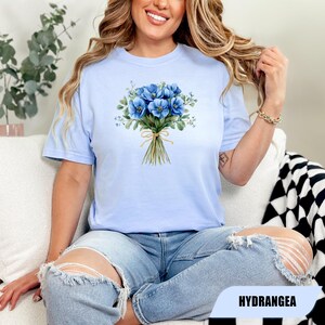 Może przedstawiać: Jasnoniebieska koszulka z akwarelowym bukietem niebieskich kwiat&oacute;w, związanym beżową wstążką. Słowo "HYDRANGEA" jest wydrukowane na czarnym prostokącie. Koszulka jest noszona przez osobę.