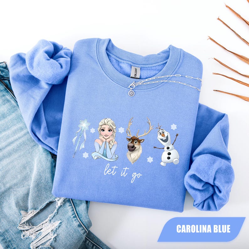 May include: Carolina blue sweatshirt featuring Elsa, Sven, and Olaf from Frozen. The graphic includes the phrase "let it go" in a script font. A silver chain necklace is draped on the neckline. The sweatshirt is displayed on a pair of blue jeans.