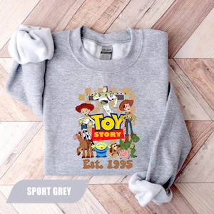 Puede incluir: Sudadera gris deportiva con un gr&aacute;fico colorido de Toy Story, con Woody, Buzz Lightyear y Jessie. El dise&ntilde;o incluye el texto "TOY STORY" en amarillo y rojo, con "Est. 1995" debajo. La sudadera est&aacute; hecha de un material suave.