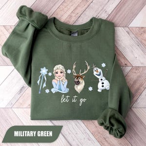 May include: Military green sweatshirt featuring Elsa, a reindeer, and Olaf from Frozen, with the phrase "let it go." The design includes snowflakes and a wand. The sweatshirt is folded on a wooden surface.