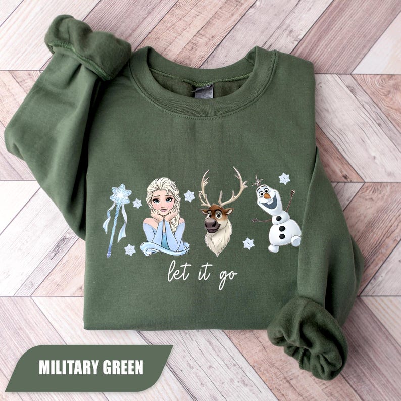 May include: Military green sweatshirt featuring Elsa, a reindeer, and Olaf from Frozen, with the phrase "let it go." The design includes snowflakes and a wand. The sweatshirt is folded on a wooden surface.