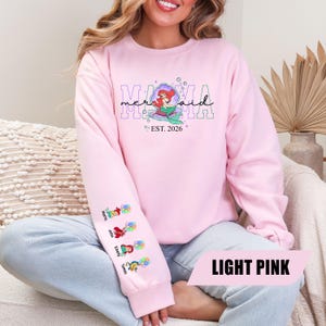 May include: Light pink sweatshirt with the text "Mama Mermaid" and an Ariel graphic. The sleeve features names and Ariel illustrations. The text "EST. 2026" is also included. The bottom right corner of the sweatshirt has the text "LIGHT PINK".