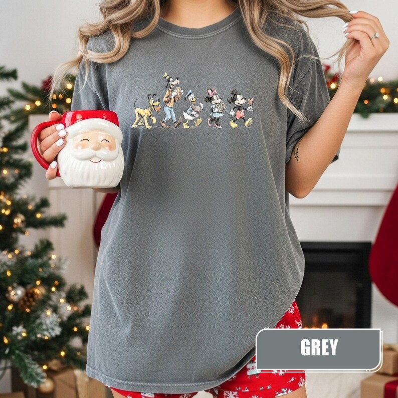 May include: A charcoal grey short-sleeve t-shirt featuring a graphic print of Pluto, Goofy, Donald Duck, Minnie Mouse, and Mickey Mouse walking in a line. The shirt is paired with a white and red ceramic mug shaped like a Santa Claus face.