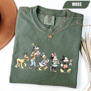 May include: A moss green crew neck t-shirt featuring a graphic print of Pluto, Goofy, Donald Duck, Minnie Mouse, and Mickey Mouse walking together while holding snacks. The shirt has a Comfort Colors brand label inside the collar.