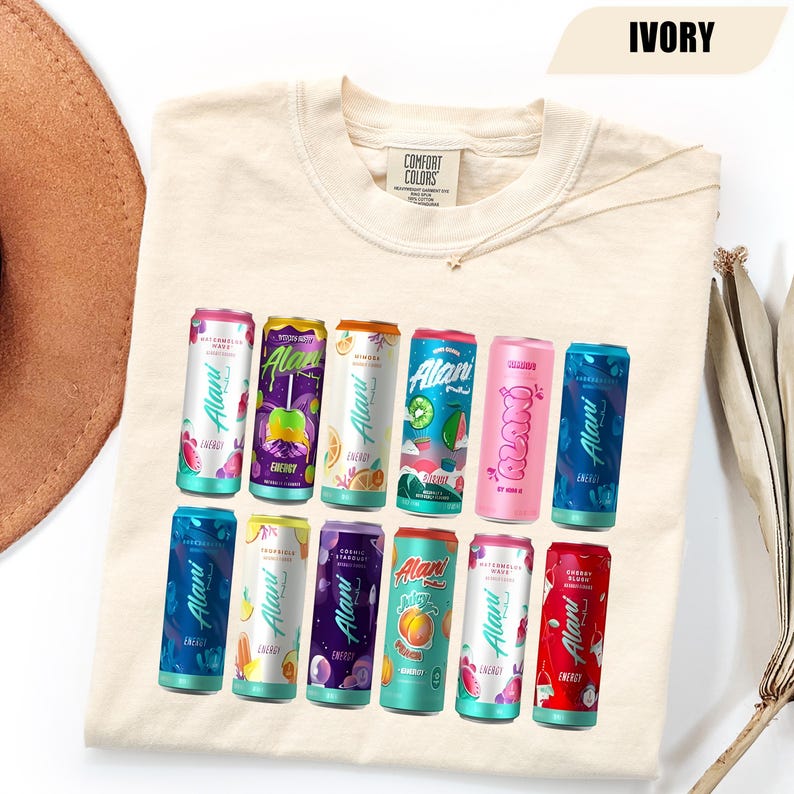 May include: An ivory crew neck t-shirt featuring a graphic print of twelve colorful Alani Nu energy drink cans arranged in two rows. The shirt has a Comfort Colors brand label at the inner neckline and the text IVORY in the top right corner.