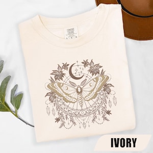 May include: An ivory t-shirt featuring a brown graphic print of a moth centered within a circular frame. The design includes a crescent moon, stars, floral elements, and hanging crystals. The neck label reads Comfort Colors with size L-G-G.