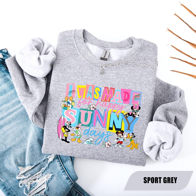 May include: A gray sweatshirt with the text "I WAS MADE for happy SUNNY days" in colorful block letters. Disney characters like Mickey Mouse, Donald Duck, and Minnie Mouse are featured. The sweatshirt is paired with blue jeans.