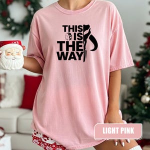 May include: A light pink short-sleeved t-shirt featuring the black text THIS IS THE WAY with a small helmet icon and a stylized silhouette graphic. The shirt has a crew neckline and a relaxed fit, showcasing a clear printed design on the front.