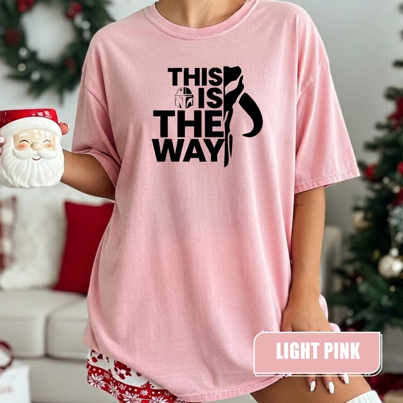 May include: A light pink short-sleeved t-shirt featuring the black text THIS IS THE WAY with a small helmet icon and a stylized silhouette graphic. The shirt has a crew neckline and a relaxed fit, showcasing a clear printed design on the front.