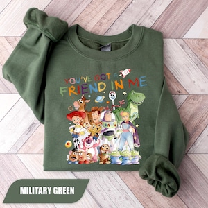 May include: A military green crewneck sweatshirt featuring a colorful graphic of various animated toy characters. The text "YOU'VE GOT A FRIEND IN ME" is printed above the characters in a multi-colored, distressed font, accompanied by small space-themed icons.