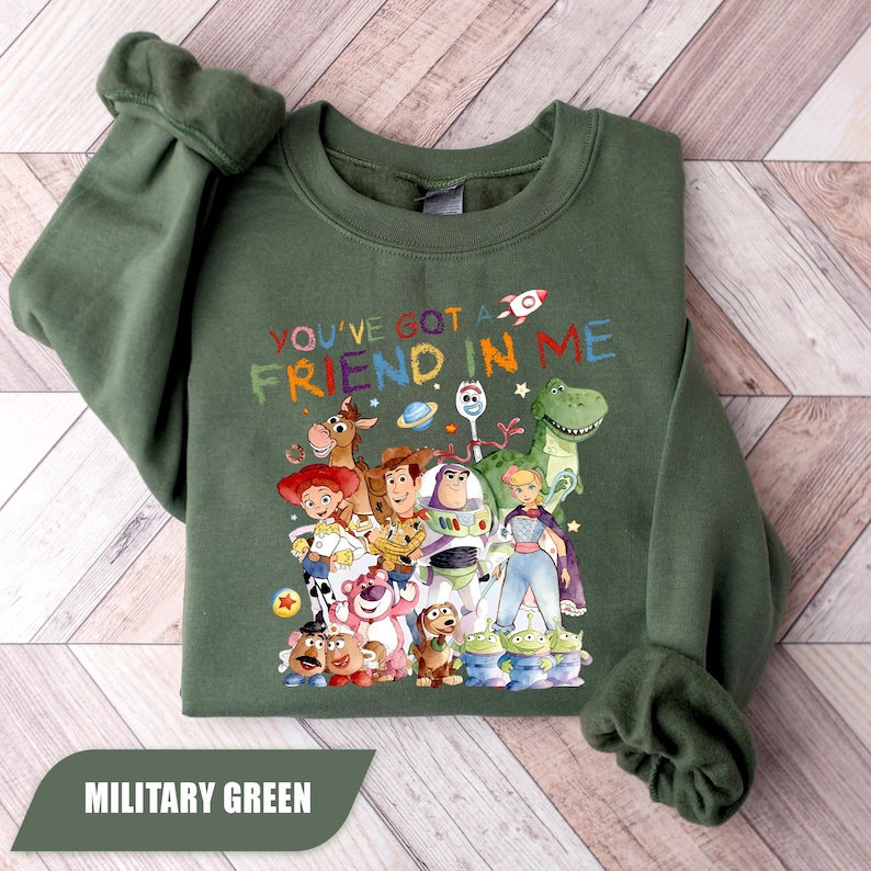 May include: A military green crewneck sweatshirt featuring a colorful graphic of various animated toy characters. The text "YOU'VE GOT A FRIEND IN ME" is printed above the characters in a multi-colored, distressed font, accompanied by small space-themed icons.