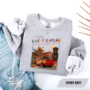 May include: A heather grey crewneck sweatshirt featuring a graphic print of a red race car on a desert road. The text reads California est. 2012, Radiator Springs, and Welcome to Radiator Springs. The design includes a water tower and rock formations.