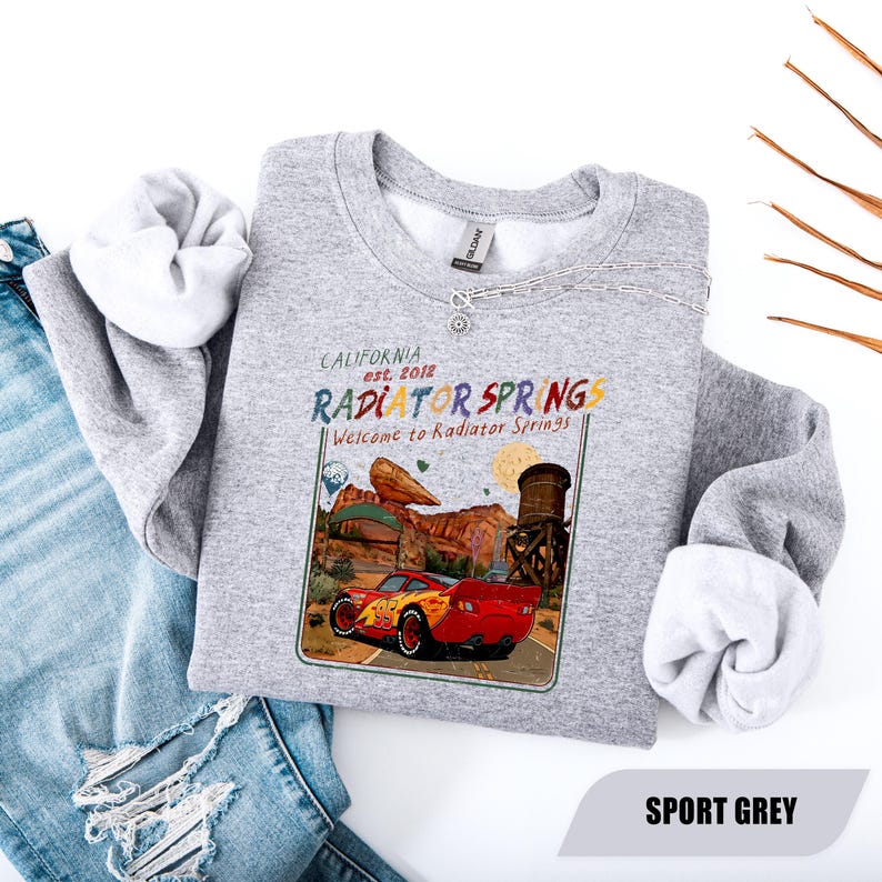 May include: A heather grey crewneck sweatshirt featuring a graphic print of a red race car on a desert road. The text reads California est. 2012, Radiator Springs, and Welcome to Radiator Springs. The design includes a water tower and rock formations.