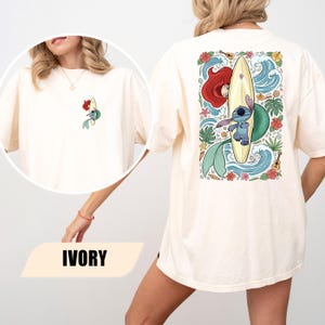 May include: An ivory t-shirt featuring a small graphic on the front left chest and a large graphic on the back. Both graphics depict a blue alien character surfing on a yellow surfboard alongside a mermaid with red hair, surrounded by tropical flowers.