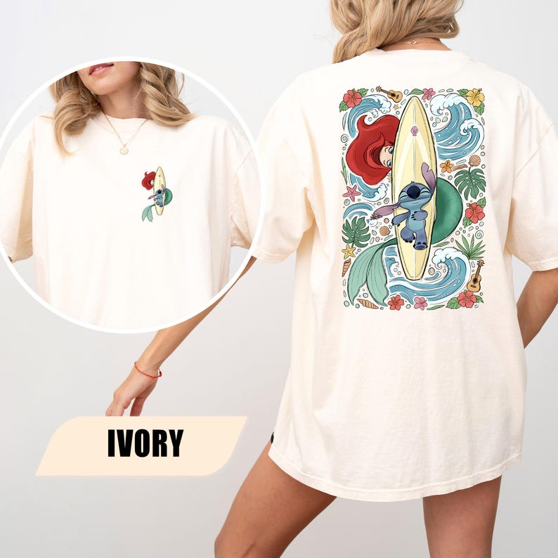 May include: An ivory t-shirt featuring a small graphic on the front left chest and a large graphic on the back. Both graphics depict a blue alien character surfing on a yellow surfboard alongside a mermaid with red hair, surrounded by tropical flowers.