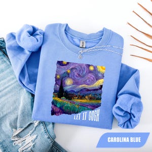 Puede incluir: Sudadera azul Carolina con un estampado de la pintura "La noche estrellada" de Van Gogh. El diseño incluye el texto "LET IT GOGH" en blanco. La sudadera se combina con un collar plateado y vaqueros azules.