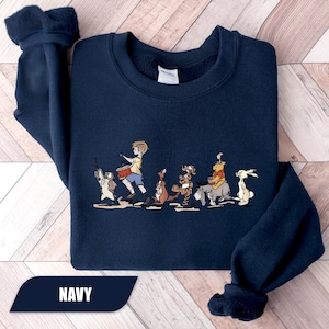 May include: Navy blue sweatshirt featuring a colorful illustration of Winnie the Pooh and friends. The graphic shows the characters marching in a line. The word "NAVY" is printed at the bottom of the sweatshirt.