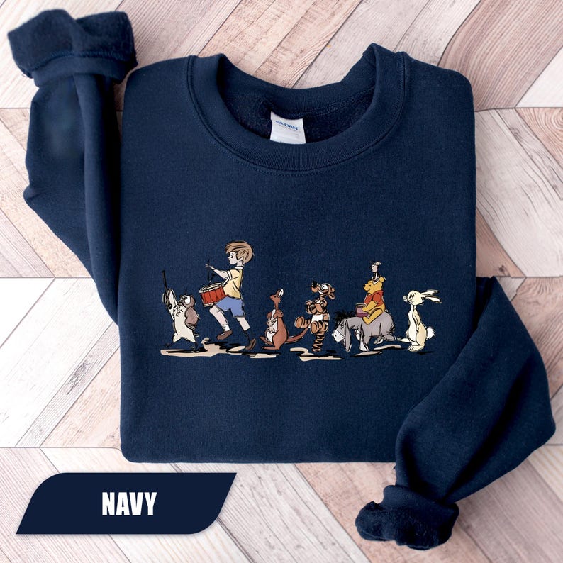 May include: Navy blue sweatshirt featuring a colorful illustration of Winnie the Pooh and friends. The graphic shows the characters marching in a line. The word "NAVY" is printed at the bottom of the sweatshirt.