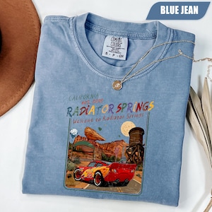 May include: A blue jean colored t-shirt featuring a graphic print of a red race car with the number 95, a desert landscape, and the text California Est. 2012 Radiator Springs Welcome to Radiator Springs. The shirt has a crew neckline and a tag.