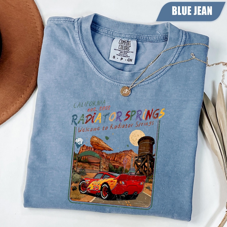 May include: A blue jean colored t-shirt featuring a graphic print of a red race car with the number 95, a desert landscape, and the text California Est. 2012 Radiator Springs Welcome to Radiator Springs. The shirt has a crew neckline and a tag.