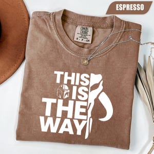 May include: An espresso-colored short-sleeve t-shirt featuring the white text "THIS IS THE WAY" alongside a graphic of a helmet. The shirt has a Comfort Colors brand label at the inner neckline, indicating a size small, and is made of 100% cotton.