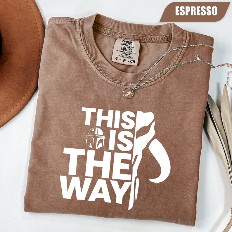 May include: An espresso-colored short-sleeve t-shirt featuring the white text "THIS IS THE WAY" alongside a graphic of a helmet. The shirt has a Comfort Colors brand label at the inner neckline, indicating a size small, and is made of 100% cotton.