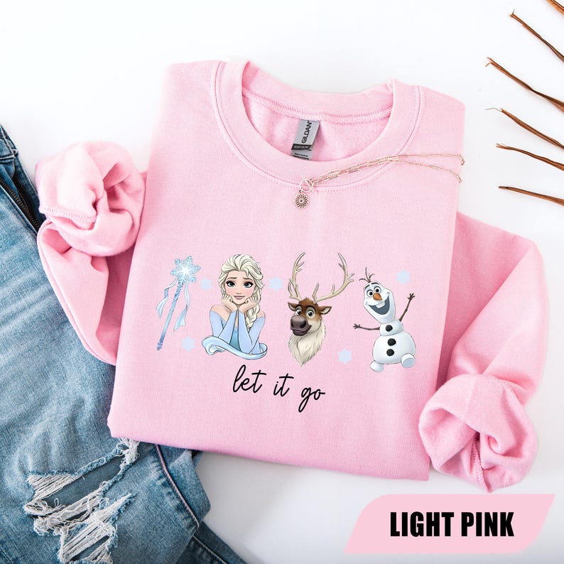May include: Light pink sweatshirt featuring Elsa, Sven, and Olaf from Frozen, with a snowflake wand and the phrase "let it go." The sweatshirt is a crew neck style.