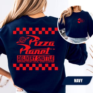 May include: A navy blue crewneck sweatshirt featuring a red graphic on the back with a checkered border, the text Pizza Planet, Delivery Shuttle, and Serving Your Local Star Cluster. A smaller red Pizza Planet logo is printed on the front left chest.