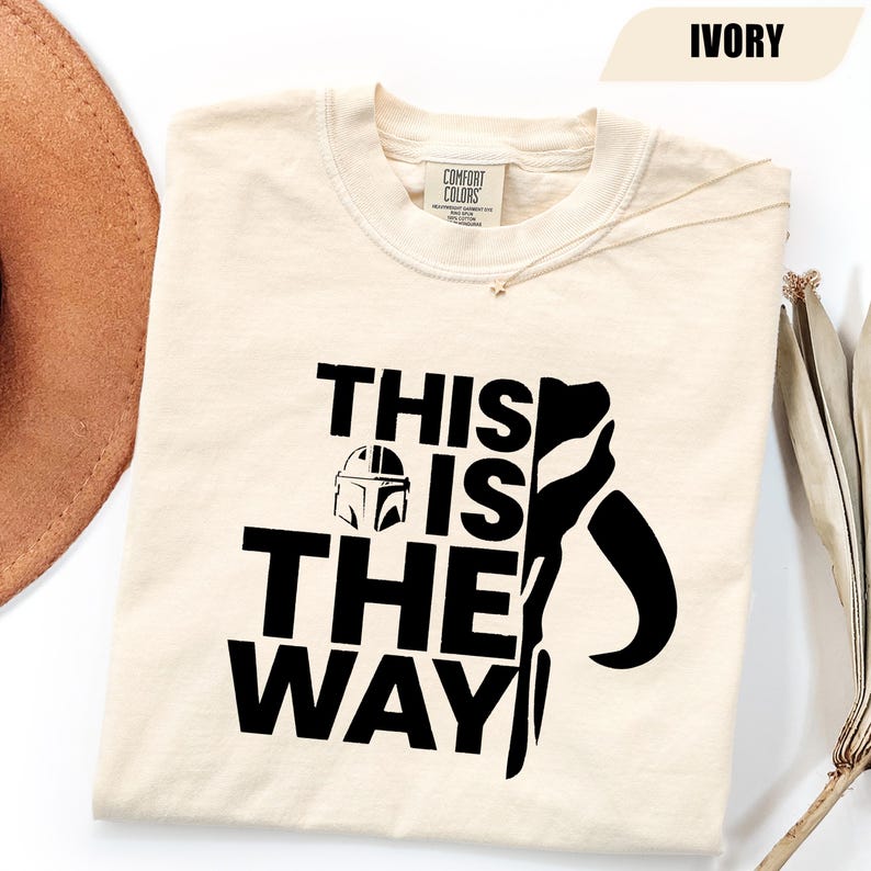 May include: An ivory-colored short-sleeve t-shirt featuring a black graphic print. The design includes the text "THIS IS THE WAY" in a bold, sans-serif font, accompanied by a small helmet icon and a large, stylized black silhouette shape.