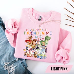 May include: A light pink crewneck sweatshirt featuring a colorful graphic print of various animated characters from the Toy Story franchise. The text "YOU'VE GOT A FRIEND IN ME" is printed above the characters in a multi-colored, playful font.