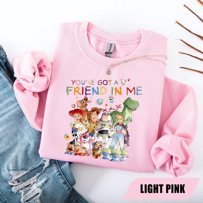 May include: A light pink crewneck sweatshirt featuring a colorful graphic print of various animated characters from the Toy Story franchise. The text "YOU'VE GOT A FRIEND IN ME" is printed above the characters in a multi-colored, playful font.