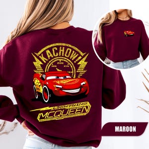 May include: A maroon long-sleeve sweatshirt featuring a large graphic on the back of the red race car Lightning McQueen with the text KACHOW and LIGHTNING MCQUEEN in yellow. A smaller version of the same red race car is printed on the front chest.